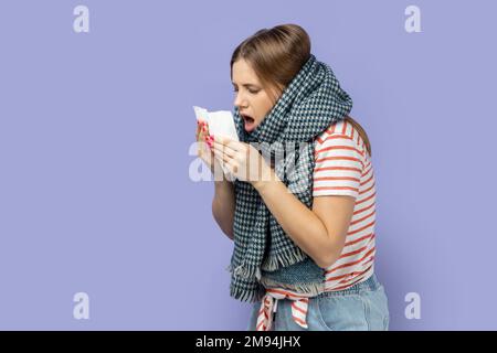 Influenza stagionale. Ritratto di donna in T-shirt a righe e avvolto in sciarpa soffiare naso che cola, starnuti in tovagliolo, che soffrono sintomi di infezione virale. Studio al coperto isolato su sfondo viola. Foto Stock