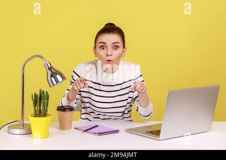 Estremamente scioccato donna lavoro in ufficio in camicia a righe seduta sul posto di lavoro, guardando e indicando la fotocamera con stupito volto sorpreso. Studio interno girato isolato su sfondo giallo. Foto Stock