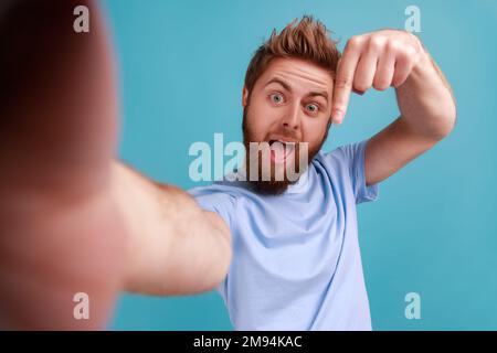 Ritratto dell'uomo bearded che scatta selfie o videochiama, guardando la fotocamera POV, il punto di vista della foto, puntando il dito verso il basso, iscriversi. Studio in interni isolato su sfondo blu. Foto Stock