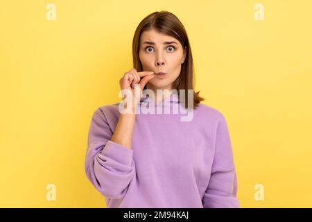 Non lo dico a nessuno. Ritratto di giovane donna adulta dai capelli scuri in piedi, guardando la macchina fotografica e zippando la bocca, indossando felpa con cappuccio viola. Studio al coperto isolato su sfondo giallo. Foto Stock