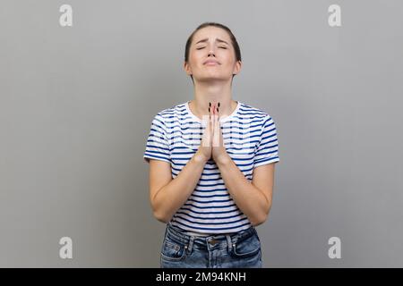 Prego, sto pregando. Ritratto di una donna preoccupata e sconvolta che indossa una T-shirt a righe che guarda in alto con implorazione disperata, pregando dio chiedendo aiuto. Studio in interni isolato su sfondo grigio Foto Stock