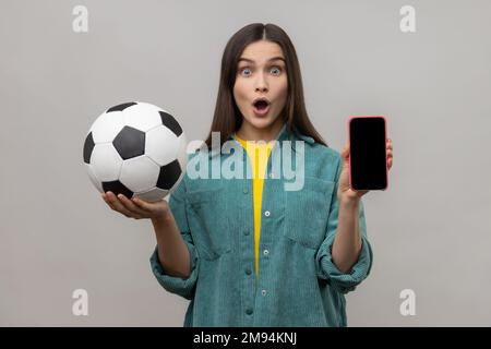 Scioccato donna sorpresa con capelli scuri tenendo e mostrando bianco display smartphone e palla di calcio, scommesse sul calcio, indossare giacca casual stile. Studio in interni isolato su sfondo grigio. Foto Stock