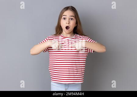 Ritratto di una bambina stupefatta che indossa una T-shirt a righe puntando su se stessa, mi chiede chi, ha sorpreso l'espressione, scioccato di essere raccolta. Studio in interni isolato su sfondo grigio. Foto Stock