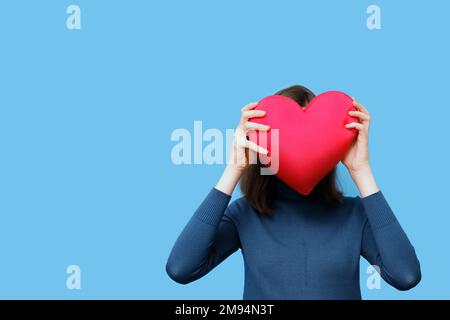 Ritratto di una bella ragazza in un maglione che tiene un cuscino rosso magenta a forma di cuore invece di un volto su uno sfondo blu. Amore, relazioni Foto Stock