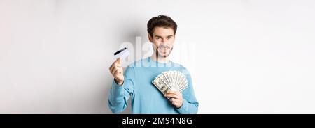 Bel giovane uomo sorridente e che offre il pagamento in contanti e senza contatto, mostrando denaro con carta di credito in plastica, in piedi su sfondo bianco Foto Stock
