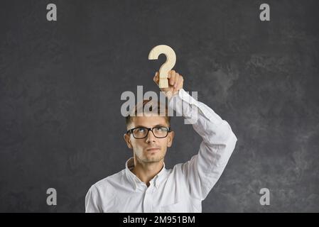 Uomo d'affari imbarazzato con emozione di dubbio sul suo volto tenendo un punto interrogativo di legno sopra la sua testa Foto Stock