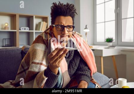 Giovane afro-americano malato che controlla la sua temperatura avvolto in una coperta a casa. Foto Stock