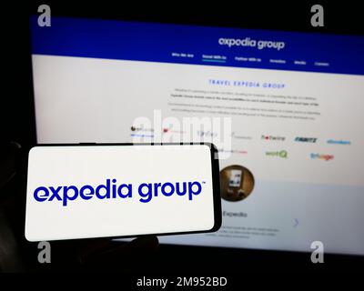 Persona in possesso di smartphone con il logo della società di viaggi online statunitense Expedia Group Inc. Sullo schermo di fronte al sito Web. Messa a fuoco sul display del telefono. Foto Stock