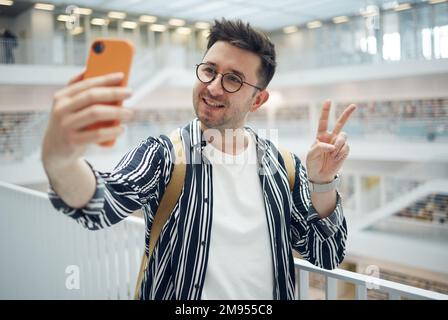 Studente universitario, uomo e libreria selfie al campus con telefono, mano di pace e sorriso sui social media. Gen z, studente universitario e app per smartphone per Foto Stock