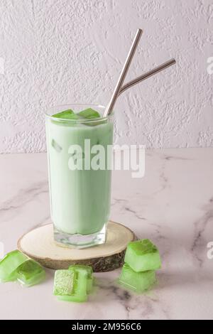 Un bicchiere alto trasparente con tè matcha verde freddo biologico e cubetti di ghiaccio, con paglia di metallo eco-friendly su un podio naturale in legno. vista frontale Foto Stock