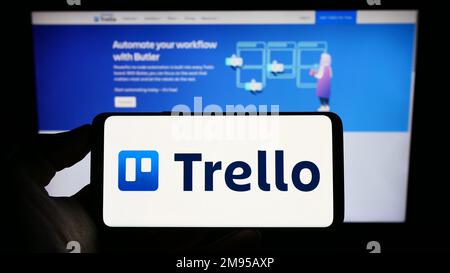 Persona che tiene il telefono cellulare con il logo del software elenco-fare Trello sullo schermo di fronte al sito web aziendale. Messa a fuoco sul display del telefono. Foto Stock