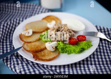 Deruny - frittelle di patate con funghi, champignons, panna acida su un piatto bianco. Pomodori ciliegini, insalata. Ristorante che serve su uno sfondo blu, tovagliolo a scacchi, forchetta, coltello. Vista dall'alto, spazio di copia Foto Stock