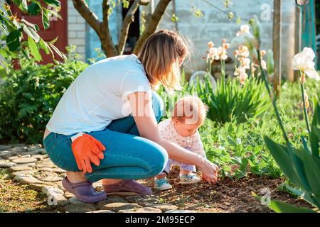 La giovane madre insegna alla figlia del bambino a curare i letti di erbe con una pala giocattolo. Vista laterale. Famiglia estate giardinaggio concetto. Foto Stock