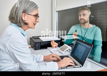 Audiologa femminile esperta che esegue test uditivi a un paziente maschio in cabina audiometrica insonorizzata con audiometro in clinica uditiva. Testo audiometrico Foto Stock