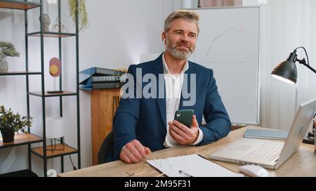 Felice rilassato overjoyed uomo d'affari maturo che lavora su computer portatile in ufficio indossando auricolari ascoltare musica da discoteca preferita energetica su smartphone. Vecchio freelance che si rilassa, facendo una pausa Foto Stock