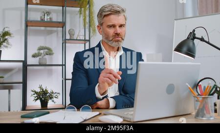 Un uomo d'affari maturo che lavora sul computer portatile scuote il dito e dice No fate attenzione a scoldarsi e dare consigli per evitare il pericolo errore disapprovazione segno in ufficio. Libero professionista sicuro uomo di mezza età Foto Stock