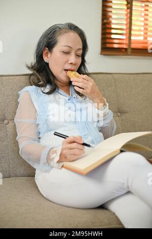 Ritratto, splendida donna asiatica di 60s anni in abiti casual mangiare biscotti e scrivere il suo diario mentre si rilassa nel suo salotto Foto Stock