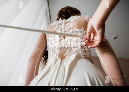 la bridesmaid aiuta a legare il nastro su un abito da sposa bianco elegante con pizzo Foto Stock
