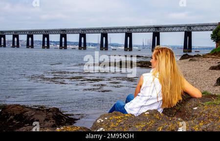 Rhianna Martin, una bella donna bionda, si siede sulle rocce ammirando la vista sulla spiaggia di Wormit nella contea di Fife, Scozia, Regno Unito Foto Stock