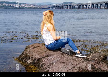 Rhianna Martin, una bella donna bionda, si siede sulle rocce ammirando la vista sulla spiaggia di Wormit nella contea di Fife, Scozia, Regno Unito Foto Stock