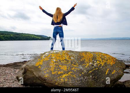 Rhianna Martin, una bella donna bionda, si erge sulle rocce mentre si gode il caldo tempo sulla spiaggia di Wormit nella contea di Fife, Scozia, Regno Unito Foto Stock
