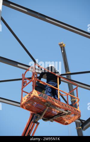 Cantiere: Lavoratore su una piattaforma di lavoro aerea che installa una struttura metallica su un edificio in costruzione Foto Stock