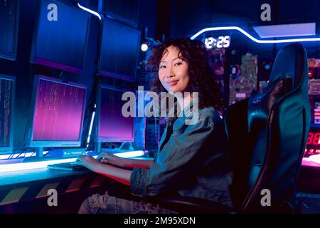 Hacker asiatico femminile sorridente in un luogo di rifugio con ambienti bui e più schermi e cavi, cercando di rompere in server di dati e infettare il sistema con un virus. Foto Stock
