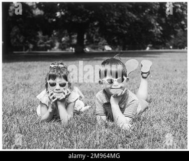 Due bambini giacciono sull'erba in un parco, ognuno con un paio di occhiali da sole. Foto Stock