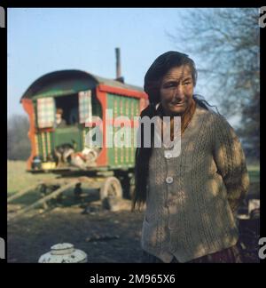 Una anziana donna zingara con lunghi capelli scuri e con uno spesso cardigan si erge di fronte alla carovana di famiglia dipinta in modo luminoso in un accampamento nel Surrey Foto Stock