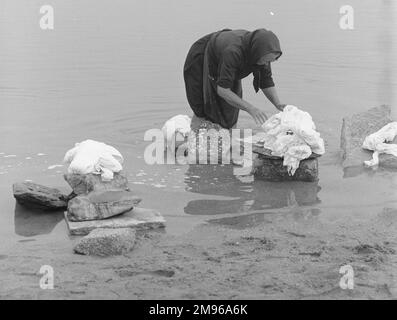 Una donna che sta in piedi ginocchio in un fiume, ordinando gli oggetti per il lavaggio che ha messo su varie rocce. Foto Stock