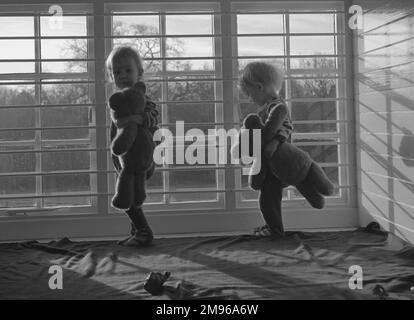 Due bambini piccoli con i loro orsacchiotti in una stanza dei bambini, in piedi davanti ad una finestra sbarrata. Foto Stock