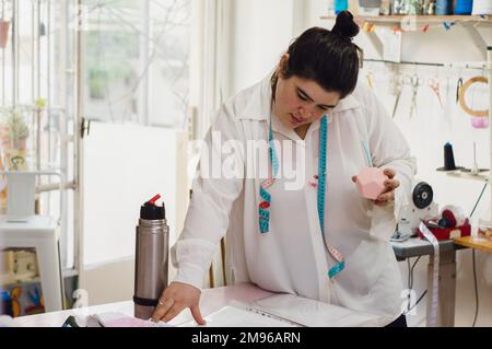 giovane argentina caucasica, con un compagno in mano, tranquillamente in piedi accanto al tavolo nel suo laboratorio di abbigliamento, guardando i disegni dei desi Foto Stock