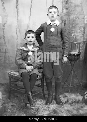 Due giovani fratelli posano per la loro foto nello studio del fotografo, probabilmente nella zona del Galles centrale. Il fratello minore è seduto su una sedia, indossando un costume da marinaio. Il fratello maggiore è in piedi su un tappeto di pelliccia, indossando una giacca Norfolk e le braghe. Foto Stock