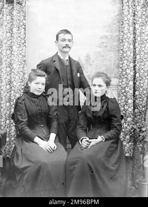 Una foto di famiglia di un giovane e di due giovani donne, possibilmente fratelli. Le donne, sedute, indossano abiti neri simili e sembrano essere in lutto. Si trovano in uno studio fotografico con uno sfondo dipinto visibile tra due tende fiorite. Foto Stock