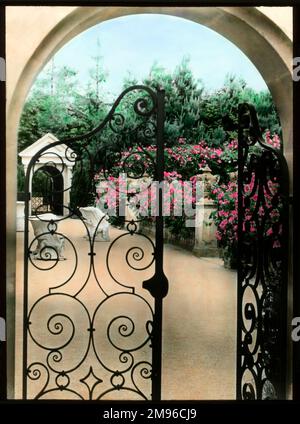 Vista sui giardini Compton Acres nel villaggio di Canford Cliffs, vicino a Poole, Dorset. Fiori rosa luminosi possono essere visti attraverso un arco con un cancello di ferro battuto. Foto Stock