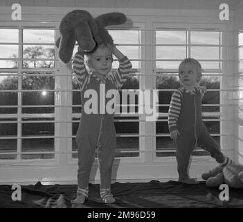 Due bambini piccoli con i loro orsacchiotti in una stanza dei bambini, in piedi davanti ad una finestra sbarrata. Foto Stock