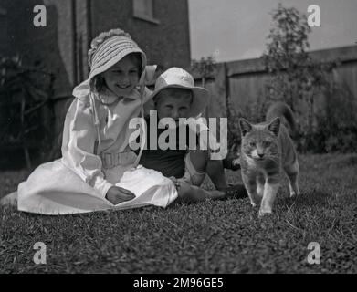 Una ragazza e un ragazzo seduti sull'erba in un giardino, a guardare un gatto. Foto Stock