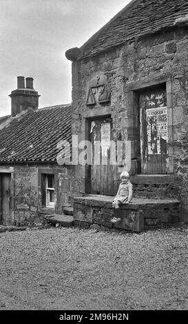 Street scene nelle Highlands scozzesi, con una bambina seduta su un gradino di pietra. Una scultura in pietra di una scala di peso porta l'iscrizione: Dio benedica i giusti. Foto Stock