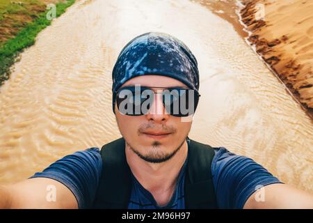 L'uomo in un'escursione con uno zaino prende un selfie sul volto della macchina fotografica in occhiali da sole e un bandana mentre viaggia su uno sfondo ruscello in Asia Foto Stock