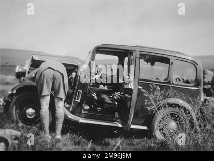 Un uomo in pantaloncini, che controlla il motore della sua auto mentre parcheggia in un campo. Foto Stock