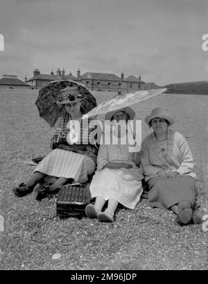 Tre donne che si siedono a terra, ombreggiandosi dal sole con due ombrelloni. Il Royal Sands Hotel sorge sullo sfondo. Foto Stock