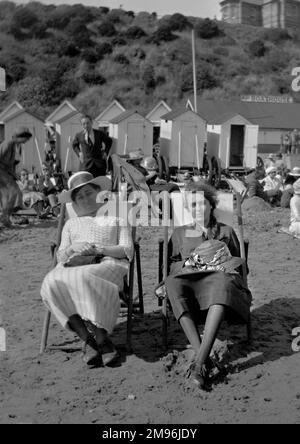 Due giovani donne sedute su una spiaggia in sdraio, con cabine sullo sfondo. Foto Stock