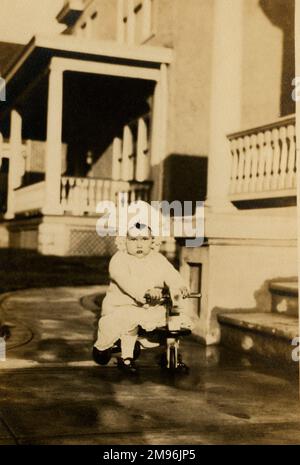 Bambino piccolo in pizzo cofano e grazioso vestito cavalca lungo il marciapiede sul suo trike di legno. Foto Stock