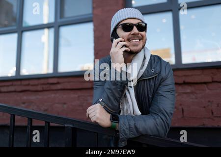 un uomo in occhiali da sole e un cappello con una sciarpa parla con un sorriso sul telefono all'esterno Foto Stock