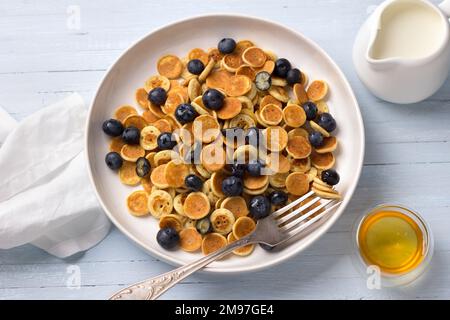 Piatto di mini pancake con cereali con crema, miele e frutti di bosco su sfondo blu chiaro. Cibo alla moda. Colazione fatta in casa. Vista dall'alto Foto Stock