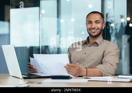 Ritratto di un giovane uomo d'affari ispanico di successo all'interno dell'ufficio, uomo sorridente e guardando la macchina fotografica, lavoratore di carta felice con i risultati di realizzazione seduto sul posto di lavoro con il laptop. Foto Stock