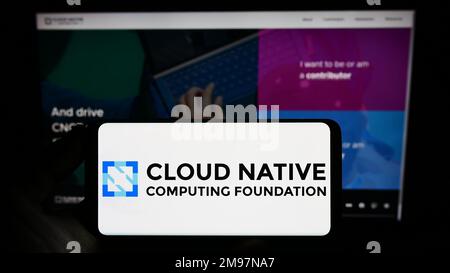 Persona che tiene in mano uno smartphone con il logo di Cloud Native Computing Foundation (CNCF) sullo schermo di fronte al sito Web. Messa a fuoco sul display del telefono. Foto Stock