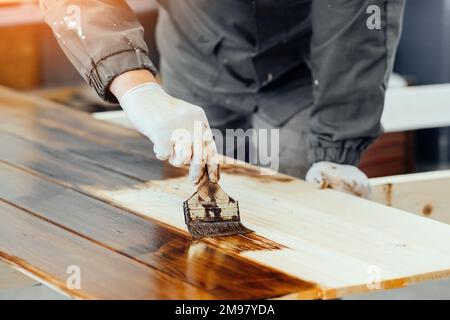 L'uomo dipinge tavole di legno con pennello. Falegname cabinetmaker vernicia la superficie di legno. Flusso di lavoro autentico. Foto Stock