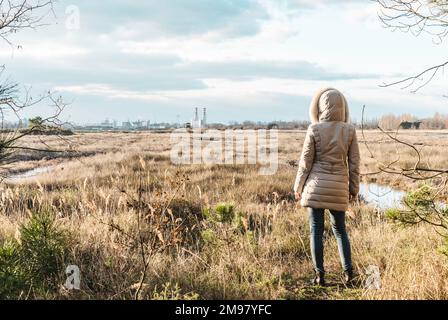 Vista posteriore della donna che indossa abiti invernali di fronte Natura selvaggia delle praterie e fabbriche all'orizzonte - paesaggio invernale con le industrie sul backgrou Foto Stock