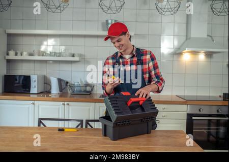 Sorridente serviceman in piedi al tavolo da cucina e guardando un paio di pinze in mano Foto Stock